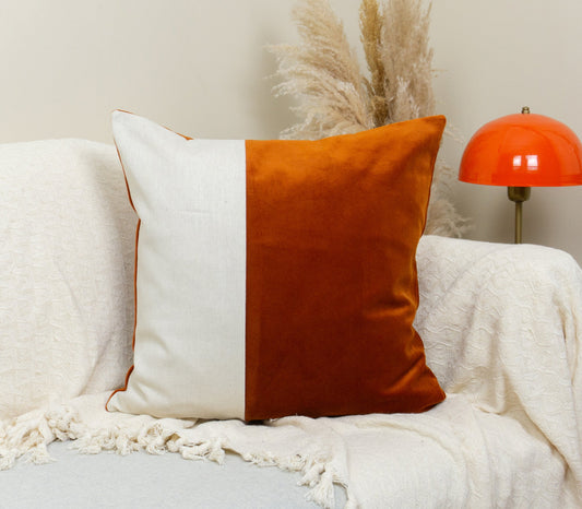 A colorblock cushion cover with burnt orange velvet and cream linen fabric displayed on a sofa with a orange-topped lamp in the background.