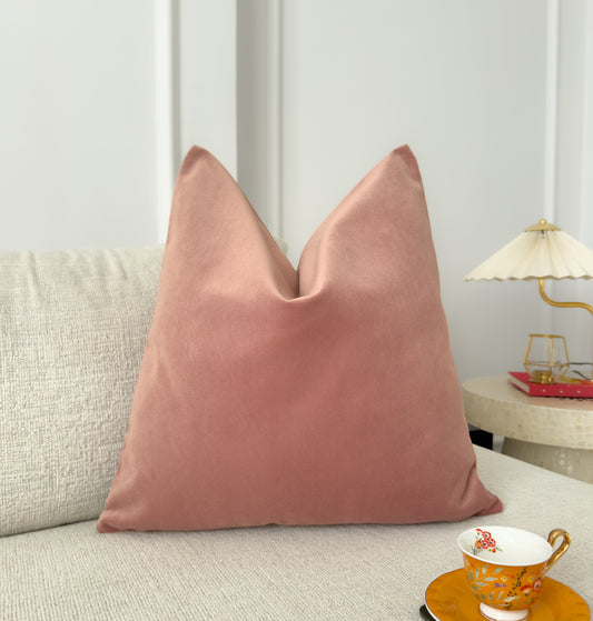 A dusty pink velvet throw pillow cover on a sofa with a plain edge, in a room setting.