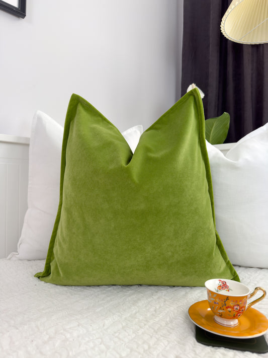 Apple green velvet cushion cover displayed on a white cushion, with a white wall and a window in the background, alongside a small orange table with a cup and saucer.